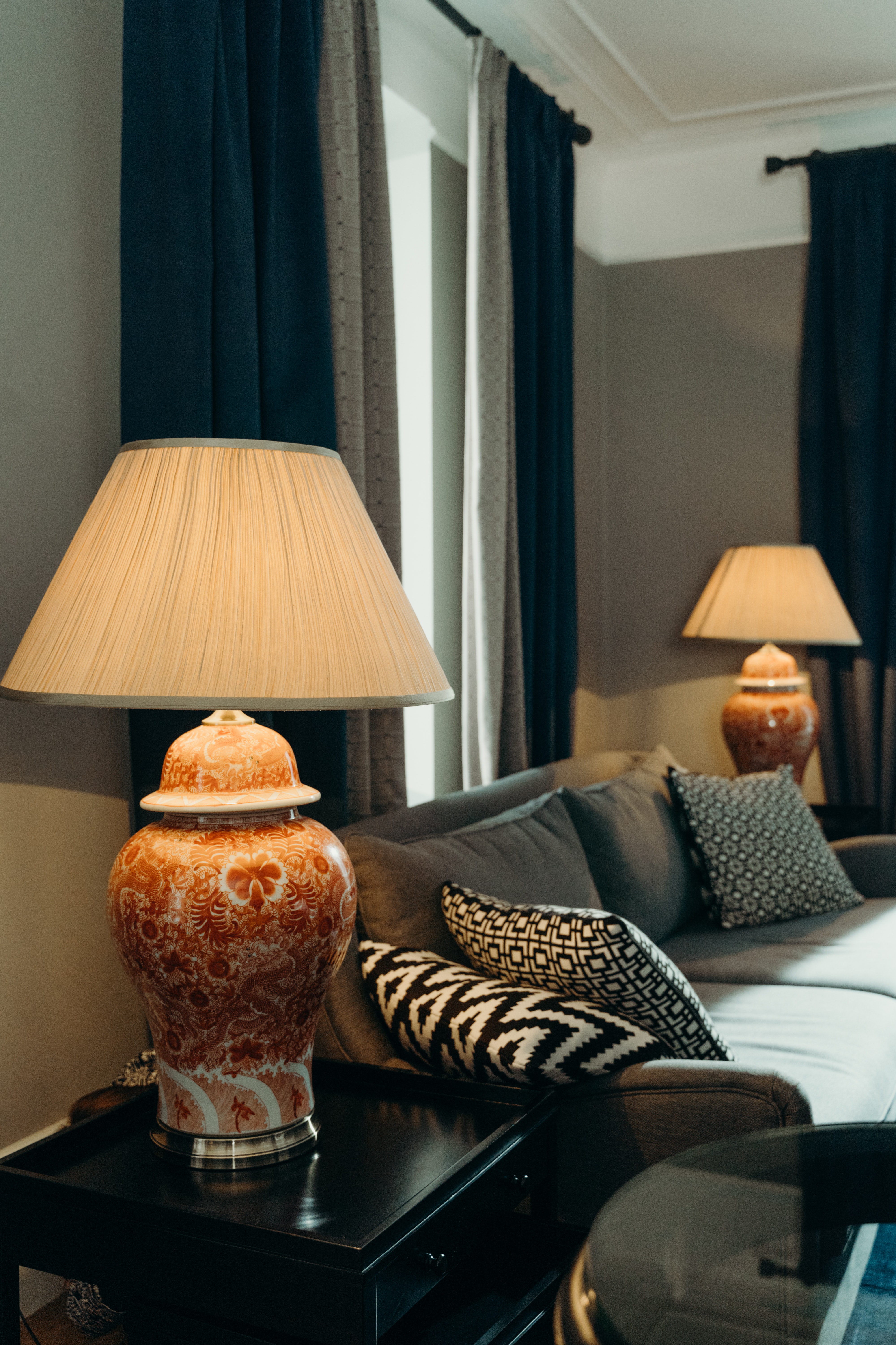 Custom-designed lampshades for antique side table lamps made of red china, and custom-designed blackout drapes in blue and silver next to a hotel couch. 
