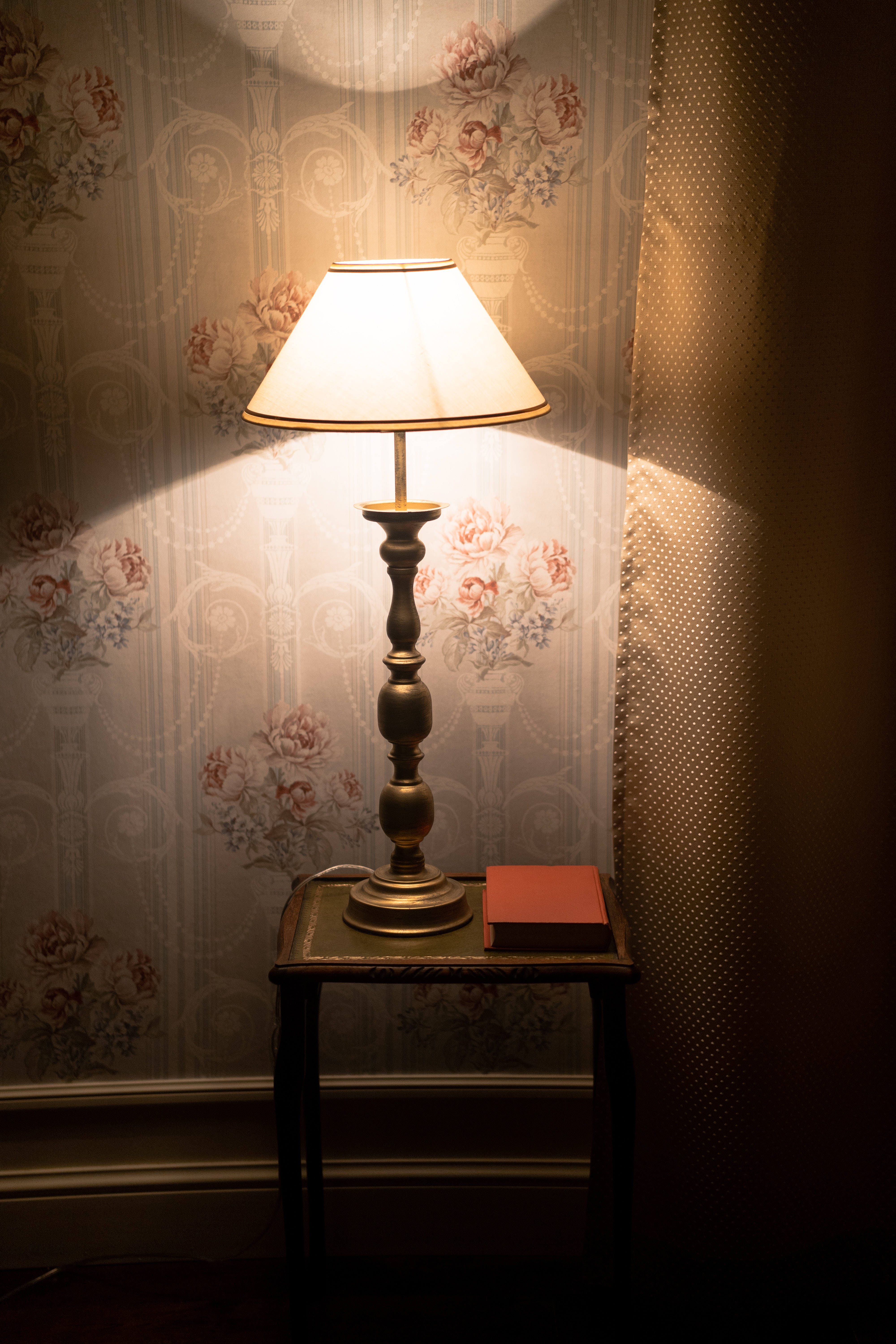 A small table lamp with a brass base and cream drum lampshade illuminates a table and wallpaper.